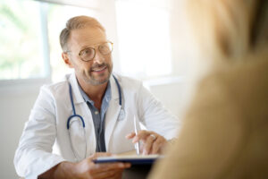 A doctor in a white lab coat smiling at a patient to the right of the frame.