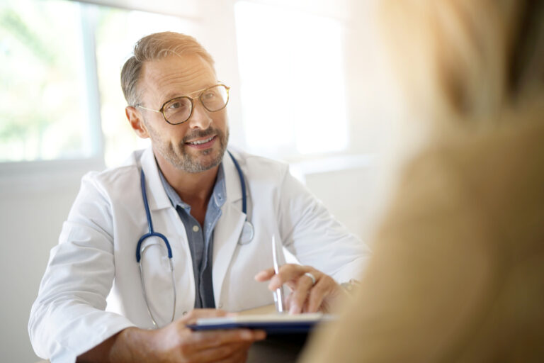 A doctor in a white lab coat smiling at a patient to the right of the frame.